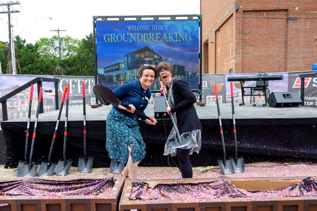 Ella Wrenn, Managing Director (left), and Mary Kate Burke, Artistic Director (right), smile and pose with shovels at the Cape Fear Regional Theatre groundbreaking ceremony. They stand in front of a confetti-filled planter and a stage with a large screen reading “Welcome to Our Groundbreaking.”